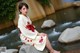 A woman in a kimono sitting on a rock by a stream.